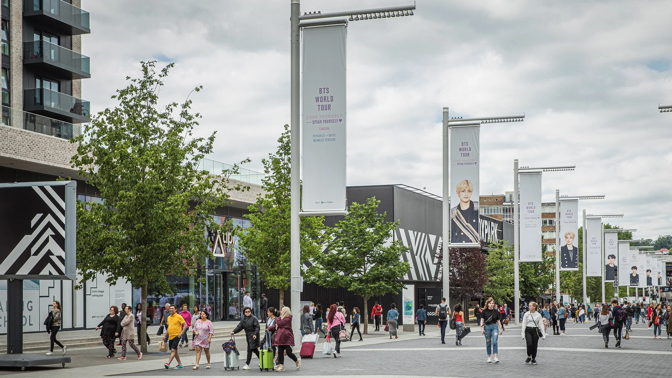 Crowd of people walking in an outdoor area with trees, banners, and a building, advertising BTS World Tour at Wembley Stadium.