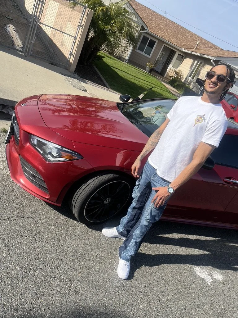 Person standing next to a red BMW car parked on a residential street with houses and a yard in the background.