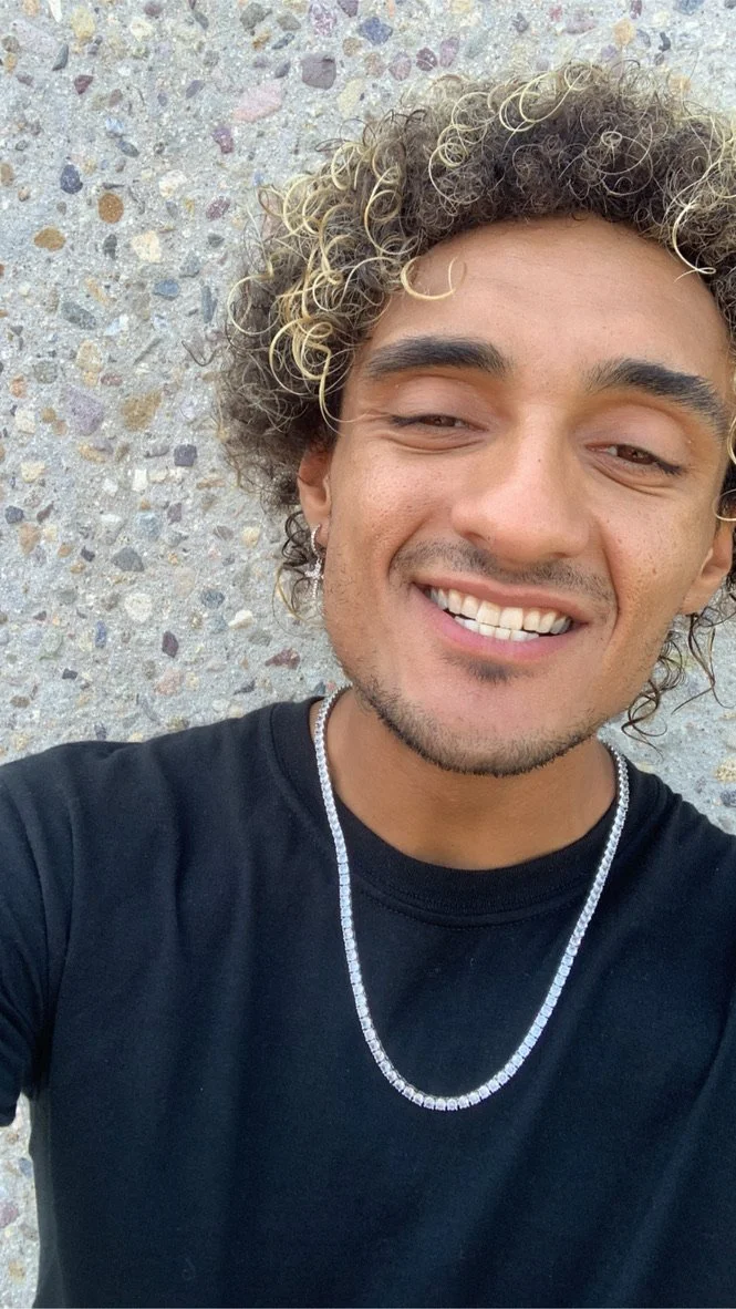 Person smiling in black shirt with a silver chain, standing against a stone wall.