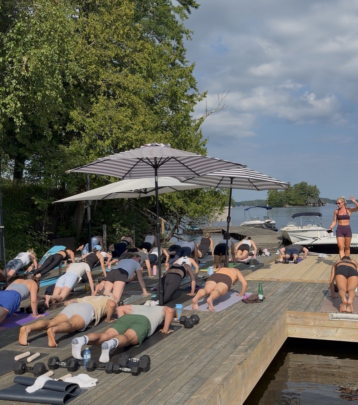 People participating in an outdoor strength class at Mortimer's Point Muskoka by the water, with boats docked nearby, under umbrellas, with fitbymich coaching and exercise gear visible.