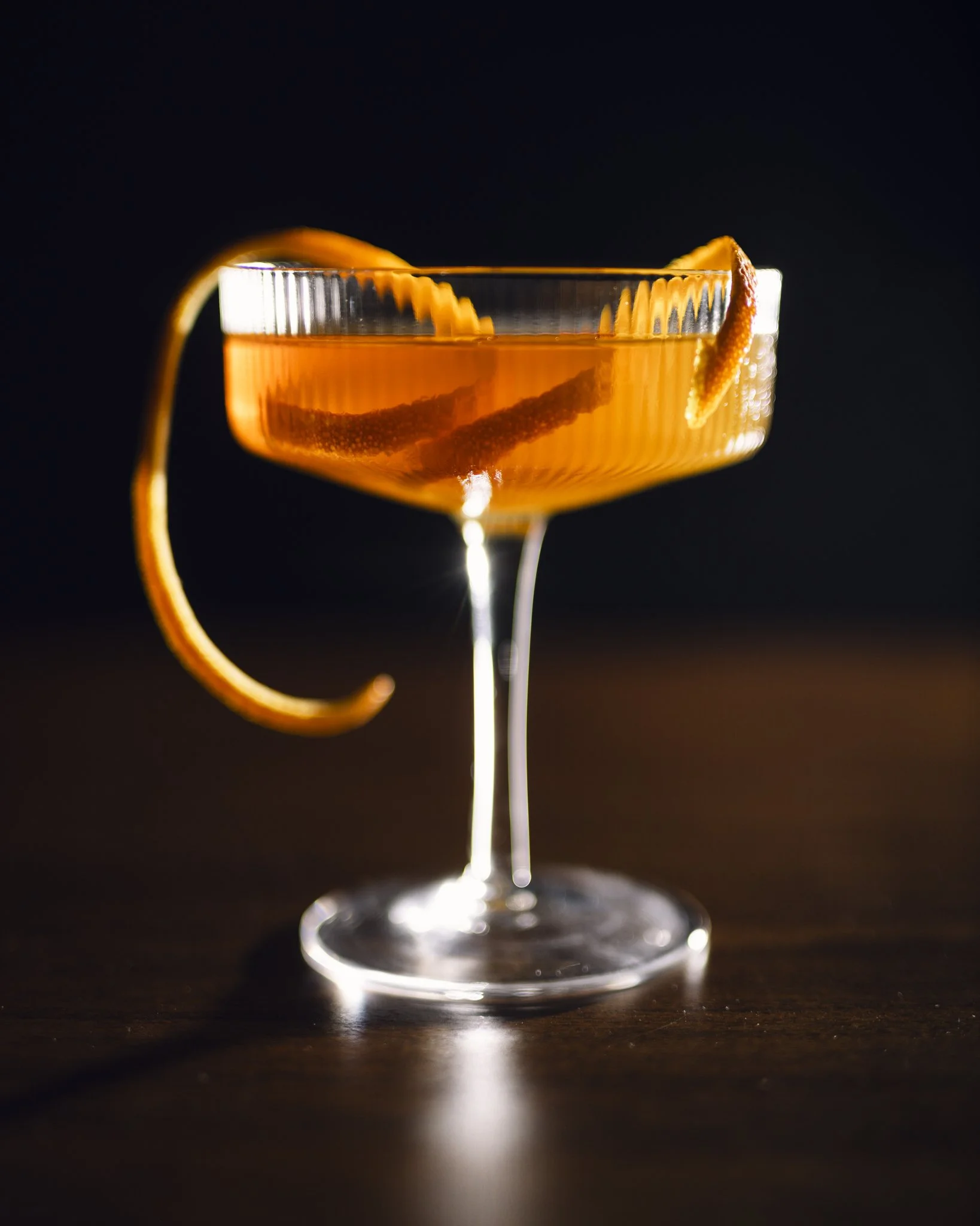 A cocktail glass with an orange-colored drink, garnished with orange peel twists, against a dark background.