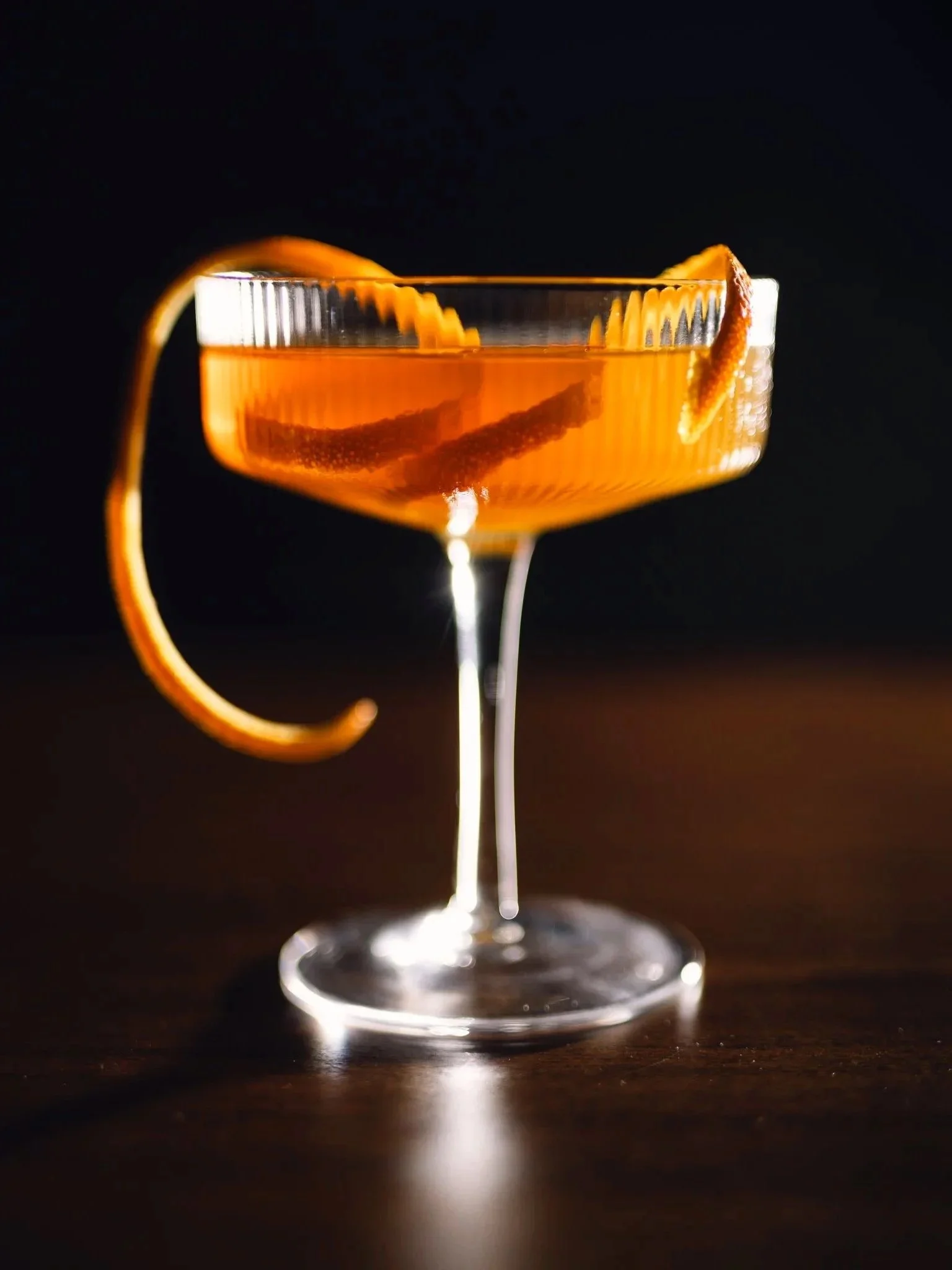 Close-up of an orange cocktail garnished with a twist of orange peel in a coupe glass on a dark background.