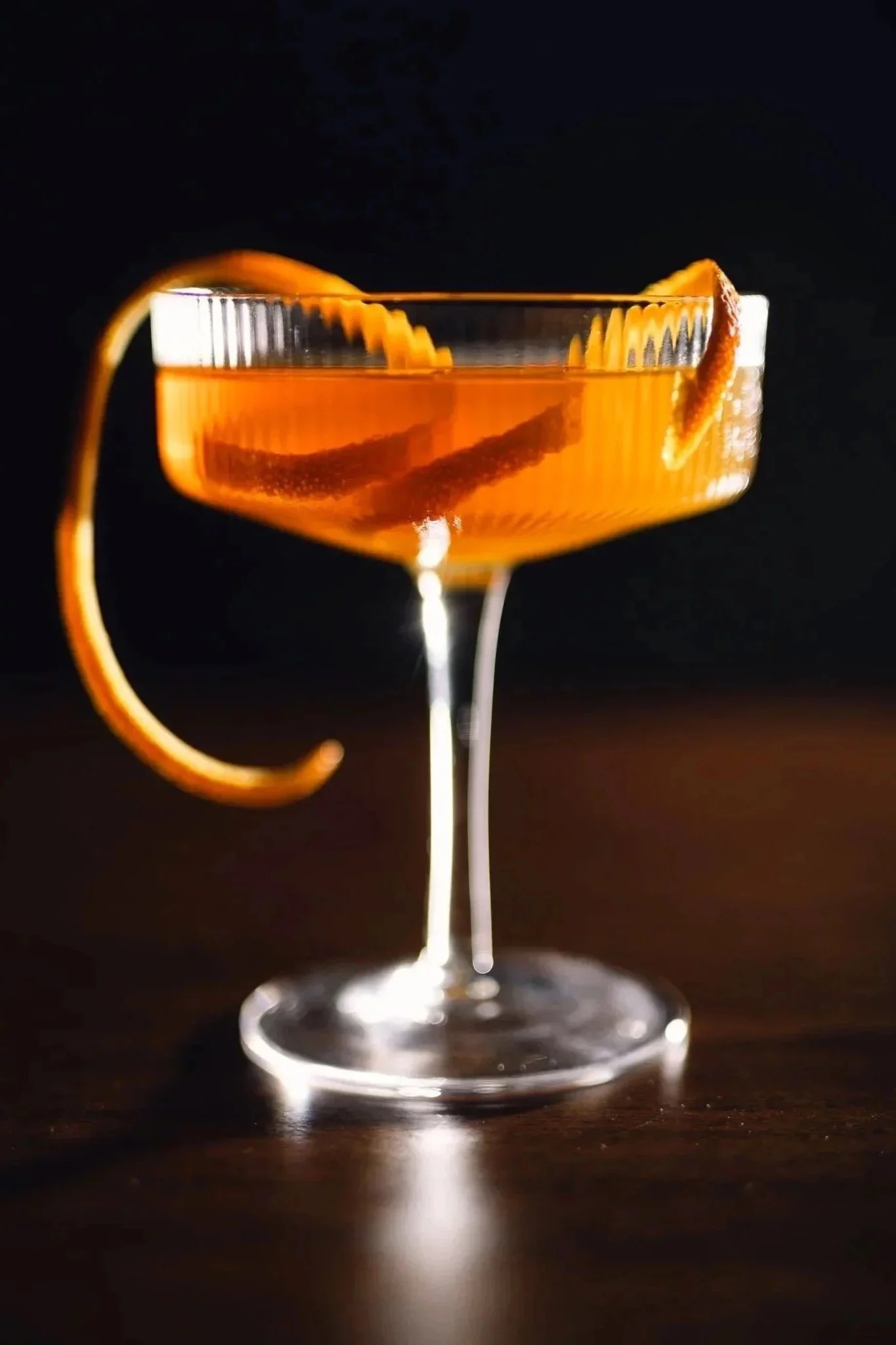 Close-up of an orange cocktail garnished with a twist of orange peel in a coupe glass on a dark background.