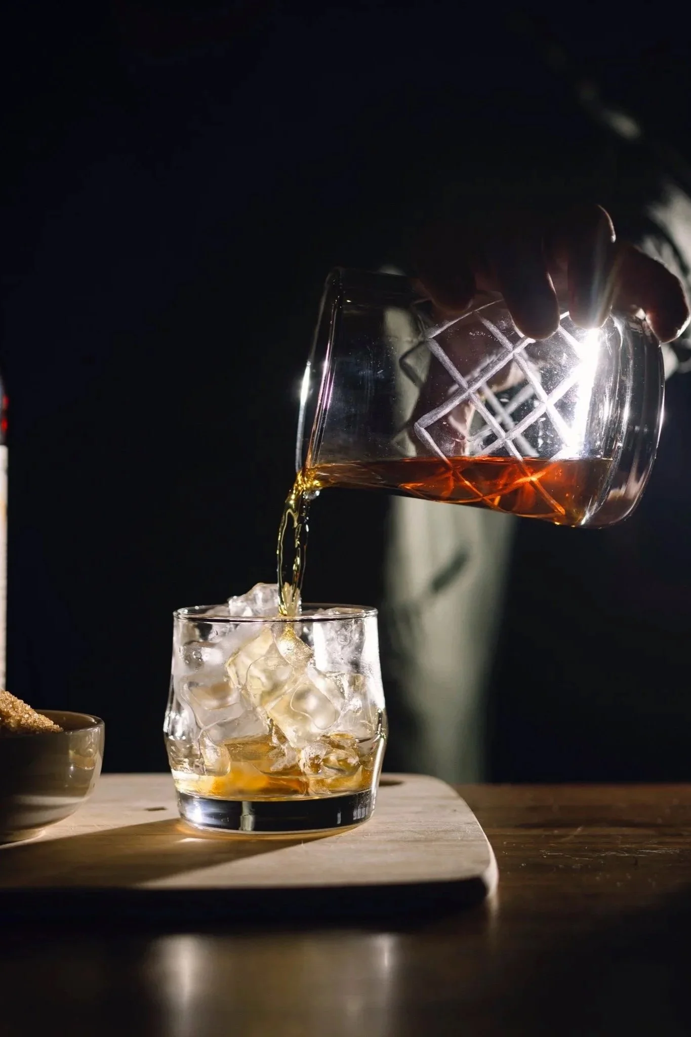 A person pours whiskey from a glass into a shorter glass filled with ice cubes on a wooden surface, with a dark background.
