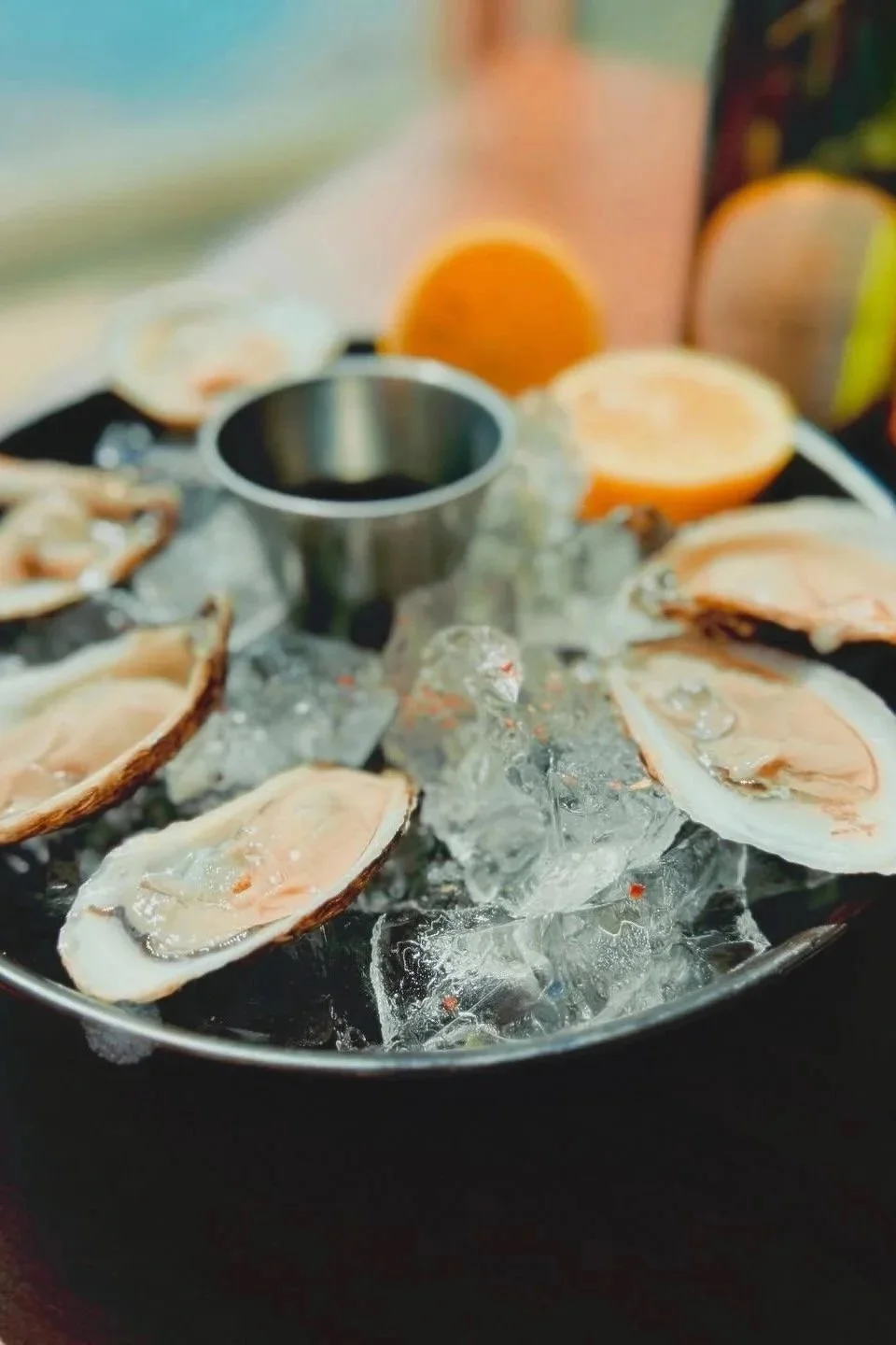 Fresh oysters on the half shell served on a bed of ice, with a small metal container of dipping sauce and sliced oranges in the background.