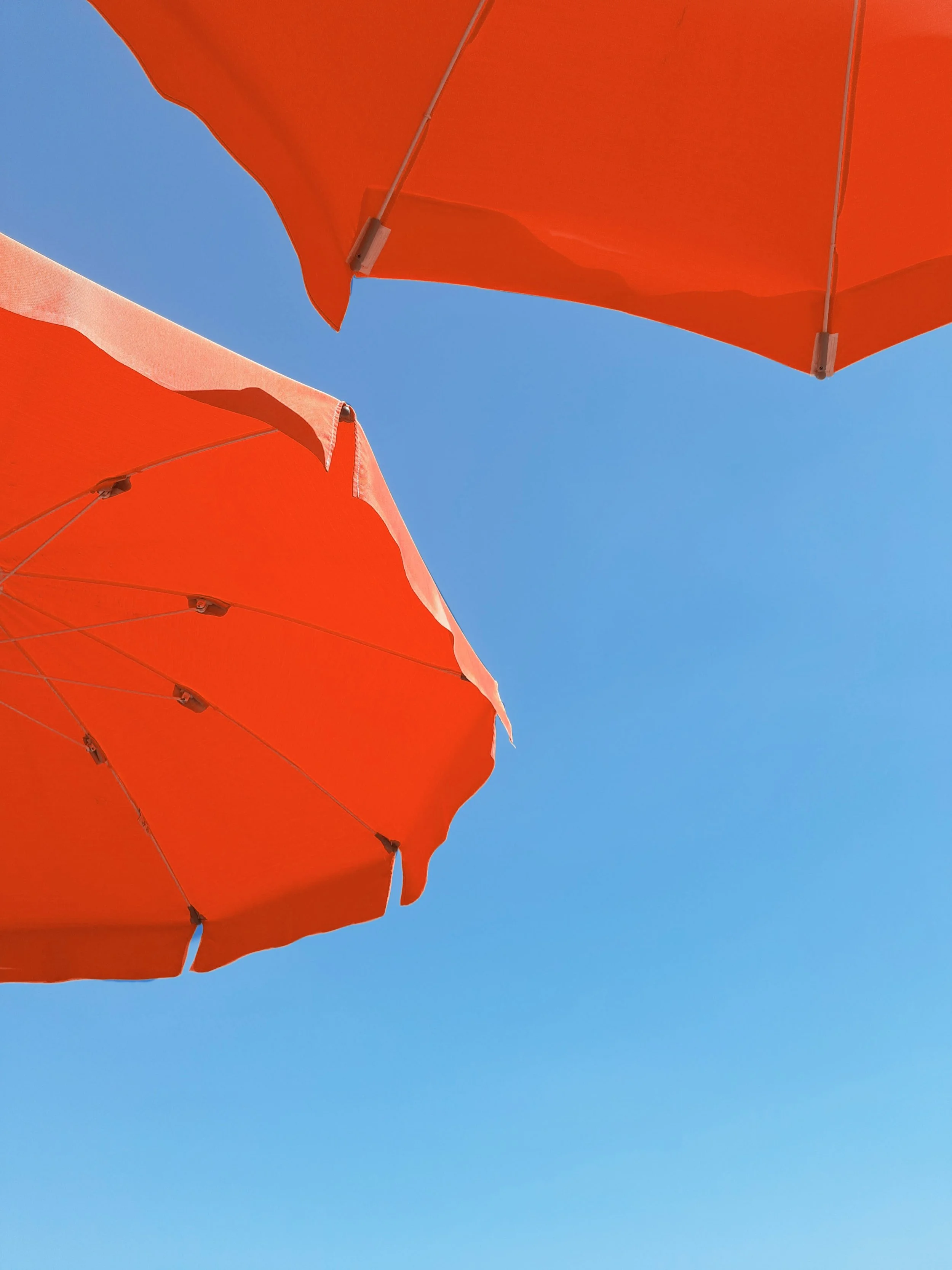 Looking up at two orange beach umbrellas against a clear blue sky.