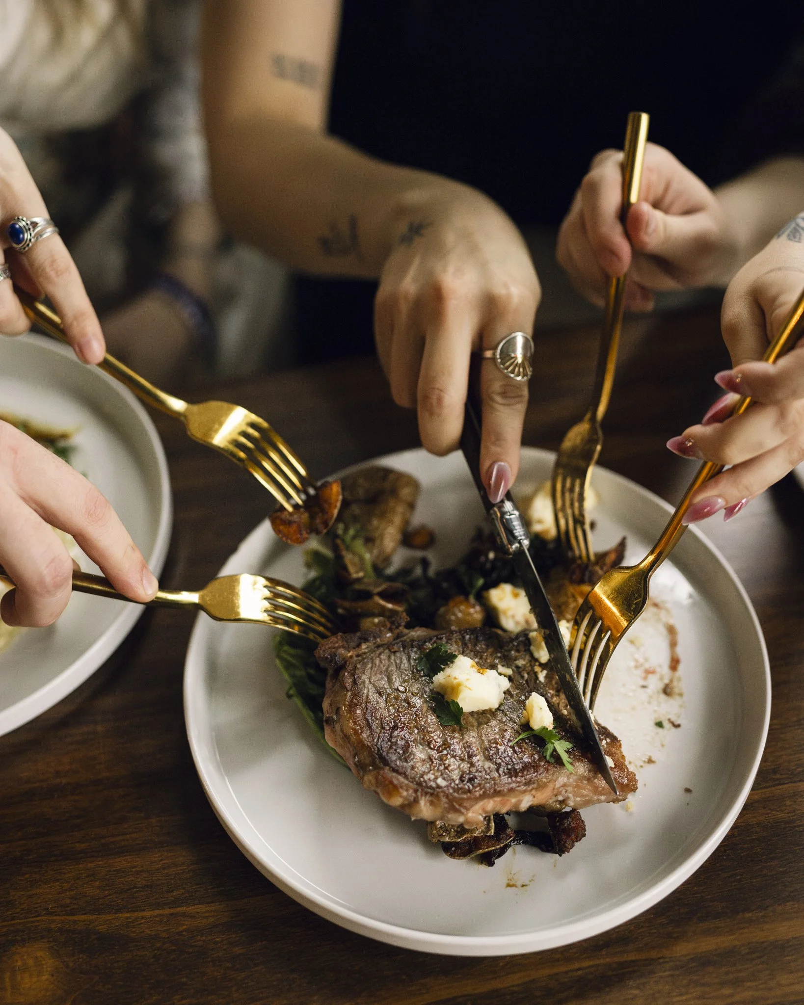 Multiple people sharing a meal, using gold forks and knives to cut into a cooked piece of steak on a white plate.