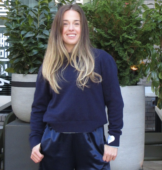 Young woman with long blonde hair wearing a navy blue sweatshirt and matching shiny navy blue pants, standing outdoors in front of large potted plants.