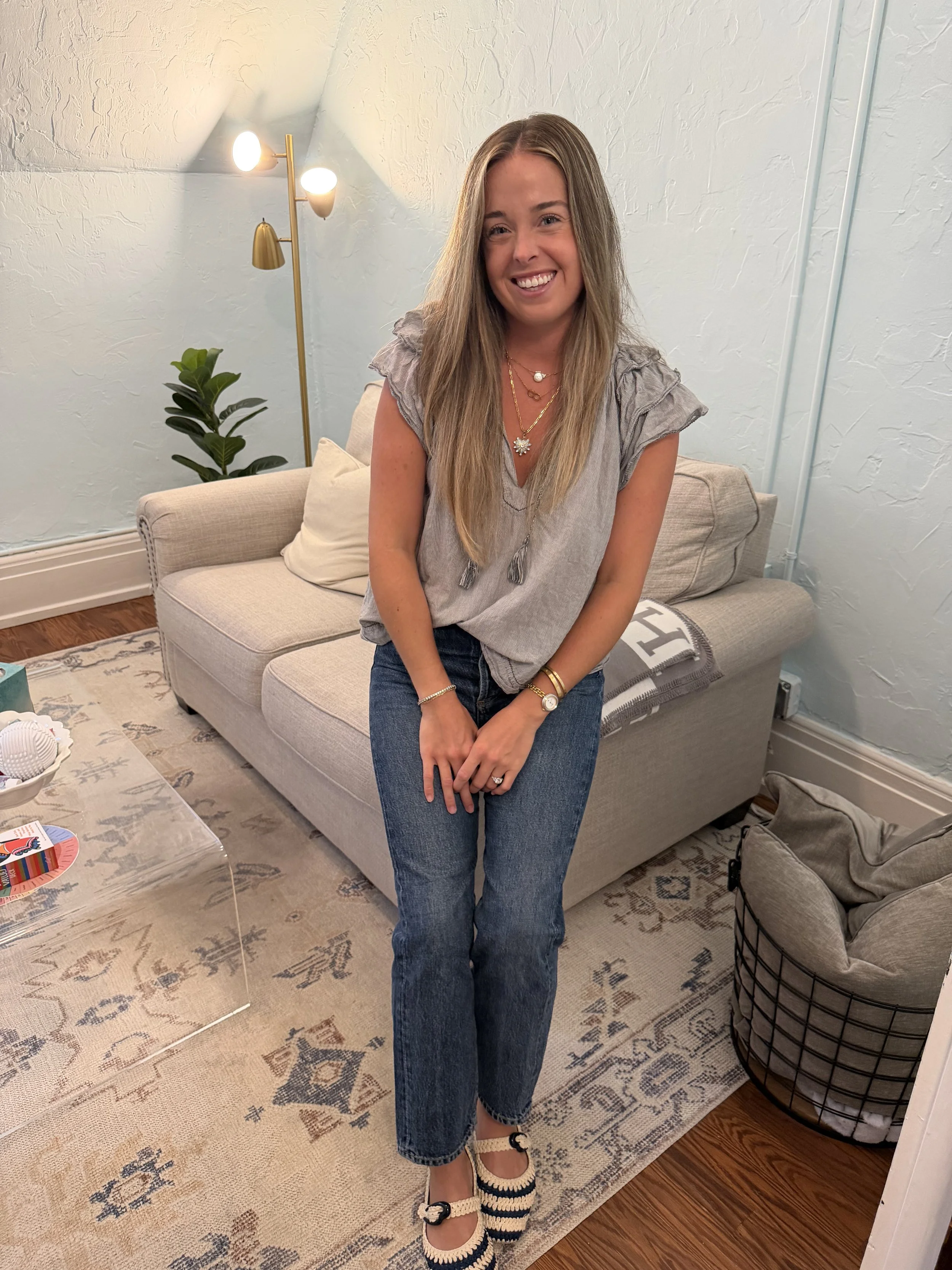 Wynne Whitley smiling in a gray blouse and jeans sitting on a light gray sofa in her therapy office.