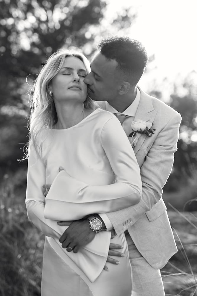 A black-and-white photo of a man and woman embracing outdoors. The man is kissing the woman's cheek, and they are both dressed in formal attire. The woman has long hair and closed eyes, while the man wears a suit with a boutonniere and a watch.