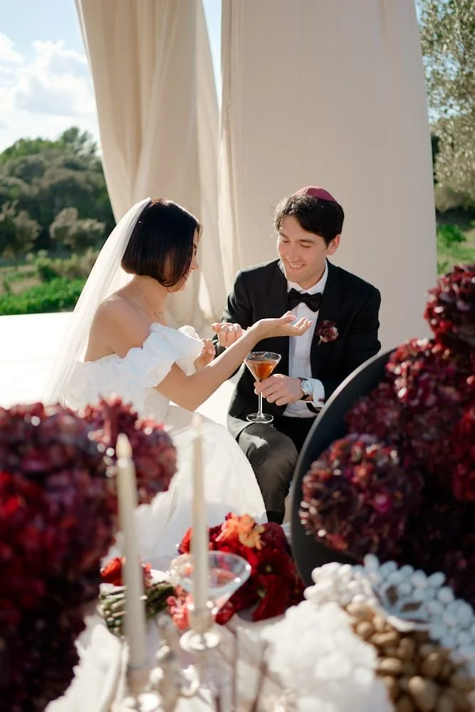 A bride and groom sharing a moment at their wedding reception, with the bride wearing a white dress and veil, and the groom in a black tuxedo with a bow tie, holding a cocktail glass, outdoors under a tent with greenery in the background.