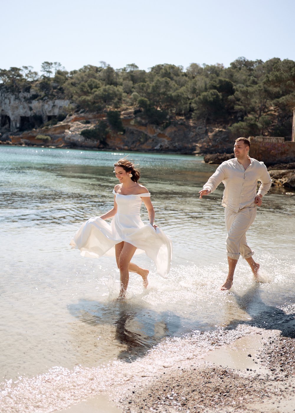 A woman in a white dress and a man in light clothing running through shallow water on a beach of Mallorca, surrounded by cliffs and trees.