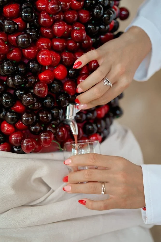 Person filling a glass with cherry juice from a bottle, surrounded by a cluster of cherries, with hands and rings visible, wearing a white long-sleeve shirt.