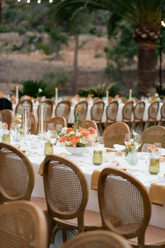 Elegant outdoor dining table set with floral centerpieces, candles, and glassware, surrounded by wicker chairs, in a garden setting.