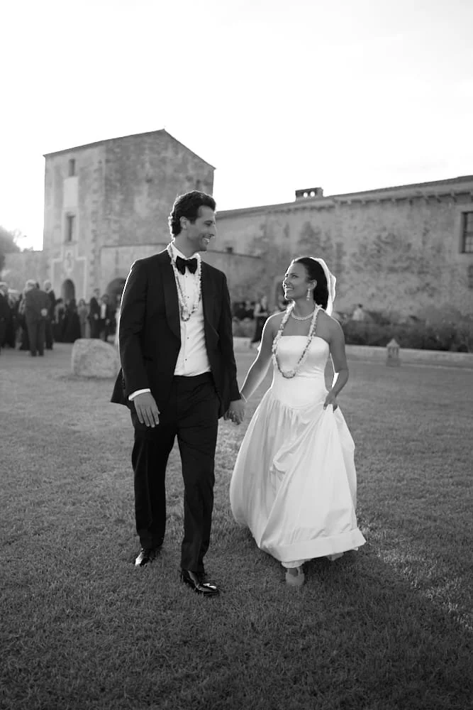 A black and white photo of a bride and groom walking outdoors on grass, holding hands, smiling at each other during their wedding. The bride wears a white strapless wedding gown with a veil and a beaded necklace. The groom wears a tuxedo with a bow t
