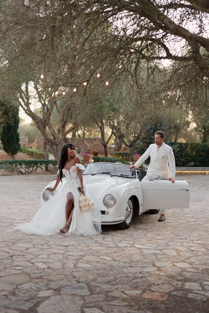 Bride in white wedding dress sitting on the hood of a vintage white convertible car, holding a bouquet, while groom in a white suit stands beside the car with the door open in an outdoor setting with trees and string lights.