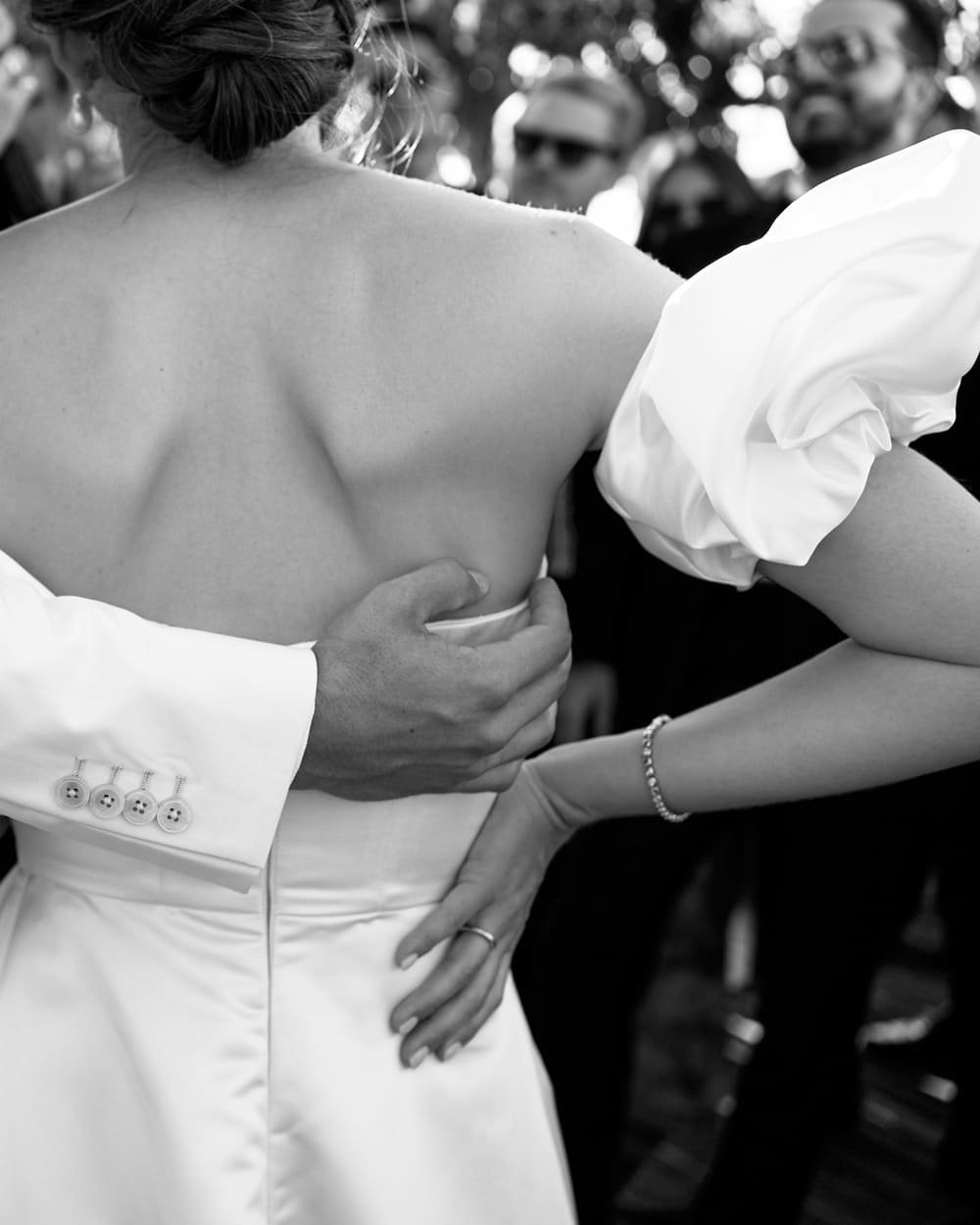 Black and white photo of a bride in a strapless dress being embraced by a person in a suit, surrounded by wedding guests.