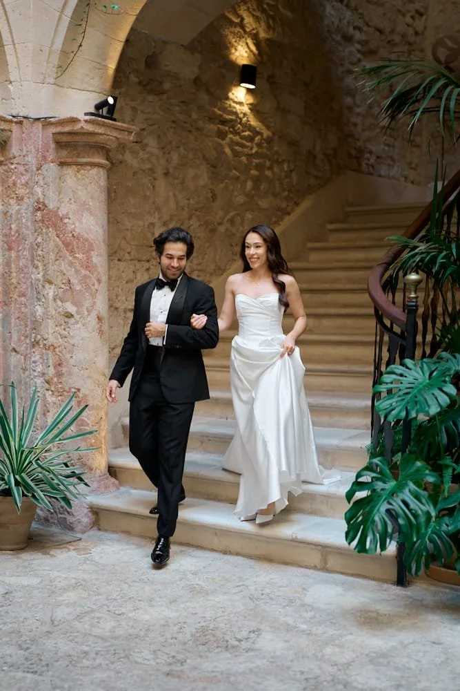 A bride and groom walking down a stone staircase inside a historic or rustic building, with stone walls and warm lighting, surrounded by potted plants.