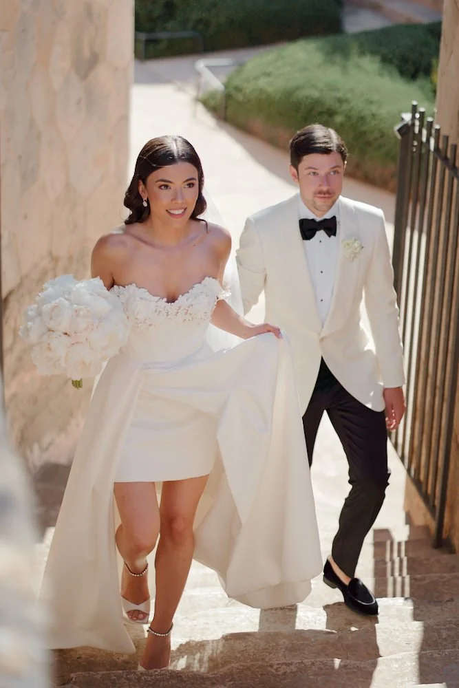 A bride and groom walking hand in hand up stone stairs. The bride is wearing a white off-shoulder wedding dress with floral detailing and holding a bouquet of white flowers. The groom is dressed in a white tuxedo jacket, black bow tie, and black pant