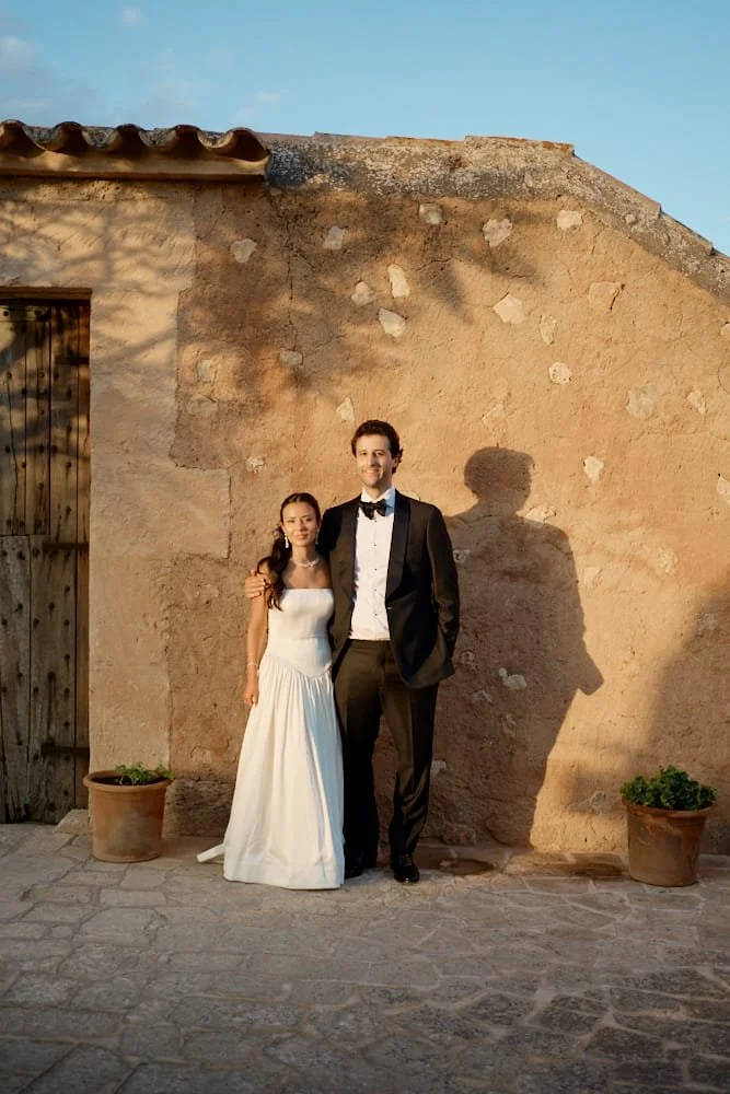A bride and groom standing together outside in front of a stone wall, with the sunset casting shadows behind them. The bride is wearing a white wedding dress, and the groom is dressed in a black tuxedo with a bow tie.
