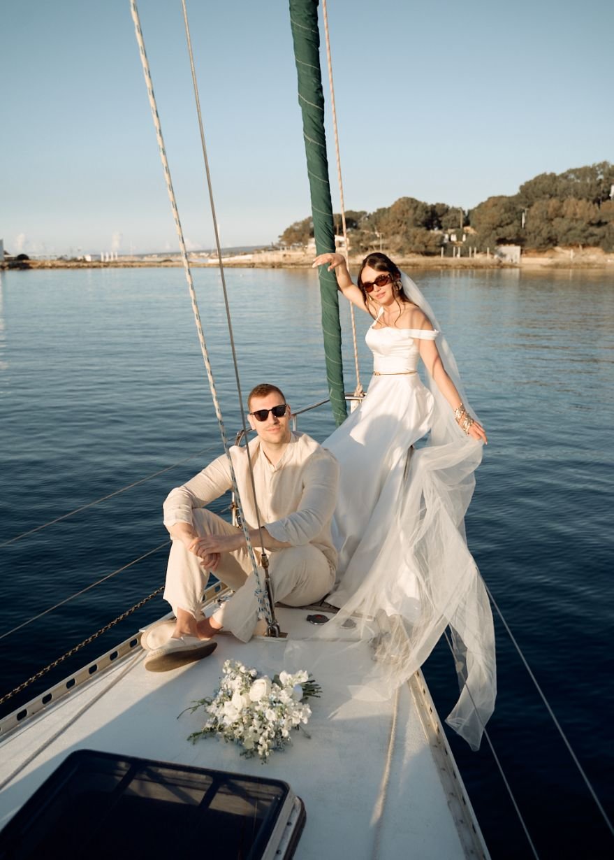 Couple in wedding attire on a sailboat; man seated and woman standing with a veil; bouquet on deck.