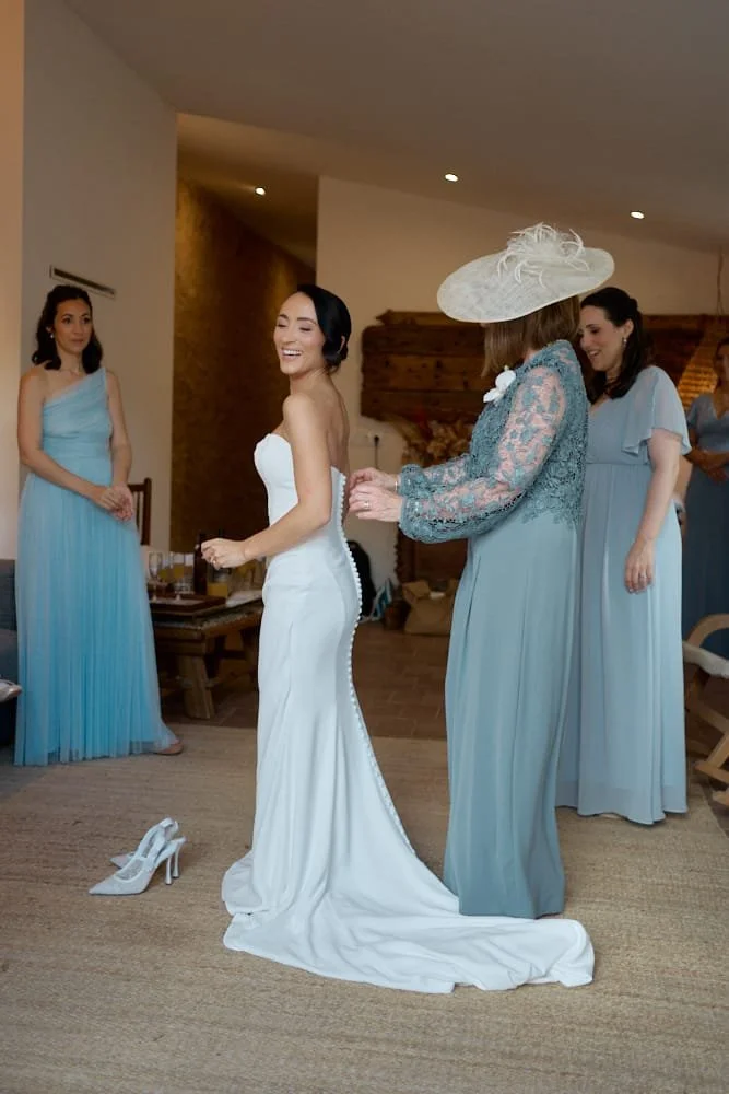 Bride in a white wedding gown smiling as she stands among her bridesmaids dressed in light blue, with a pair of white shoes on the floor nearby, in a cozy indoor setting.