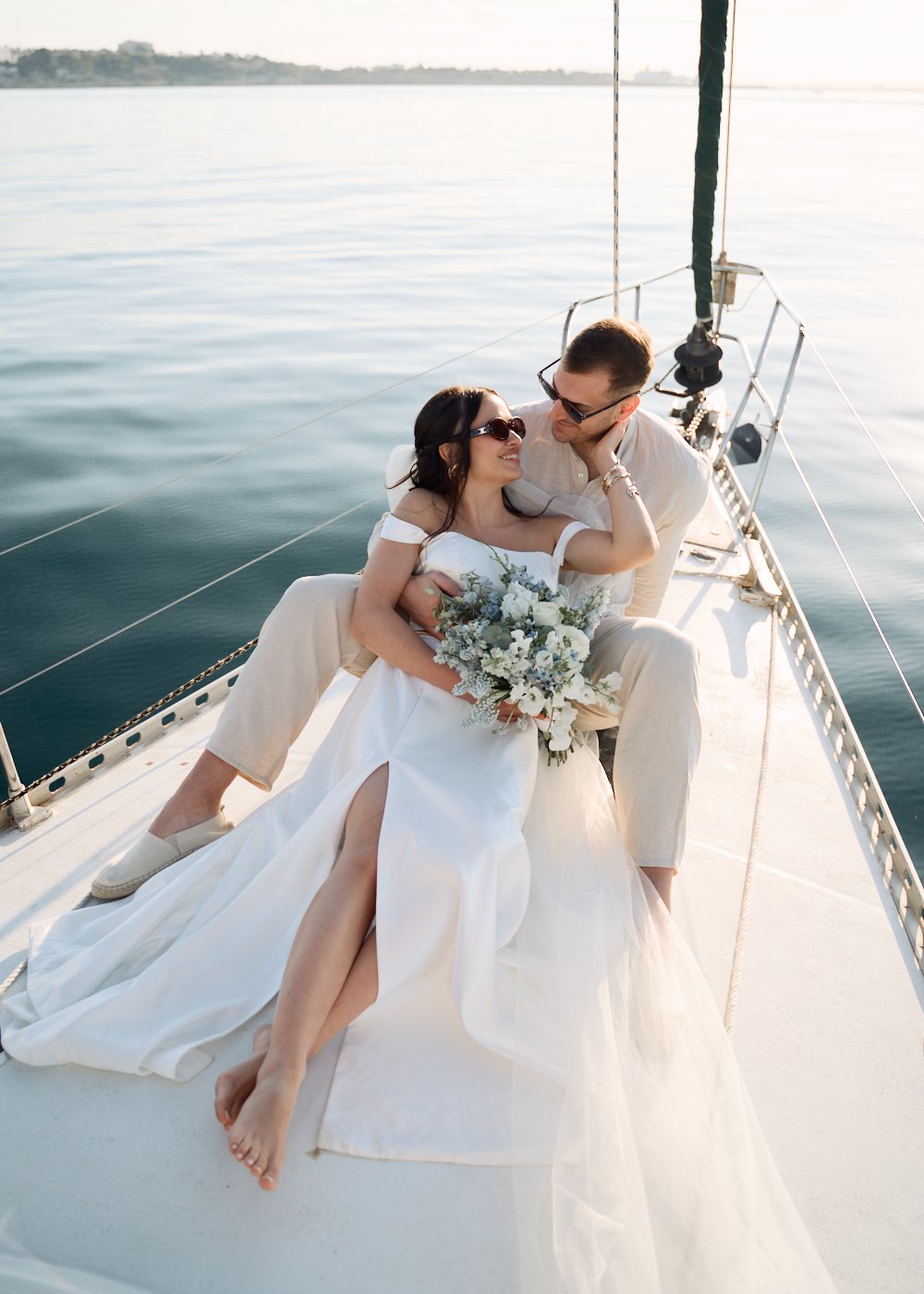 Couple in wedding attire sitting on a sailboat, with the woman holding a bouquet and both wearing sunglasses.