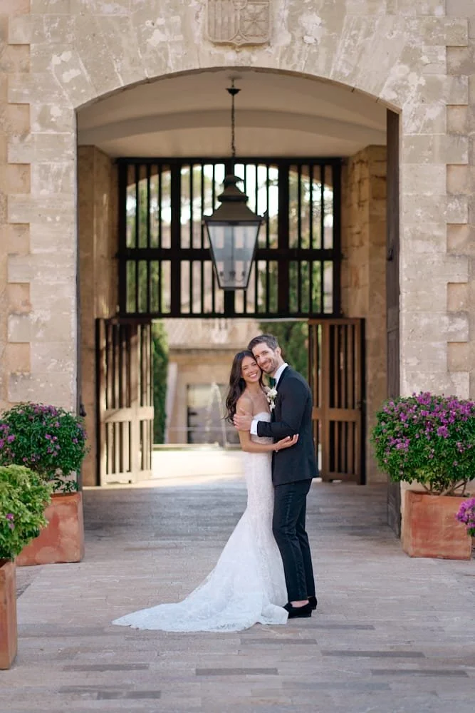 A newlywed couple hugging and smiling under a large stone archway with a lantern hanging overhead, potted purple flowers on either side, in an outdoor wedding setting.