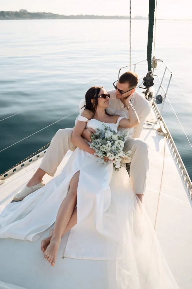 A couple dressed in wedding attire on a sailboat, with the woman holding a bouquet of flowers, smiling and looking at each other.