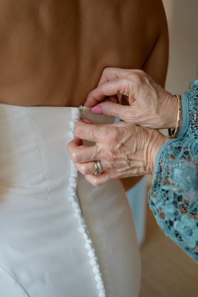 Older woman fastening a row of fabric-covered buttons on the back of a bride's wedding dress.