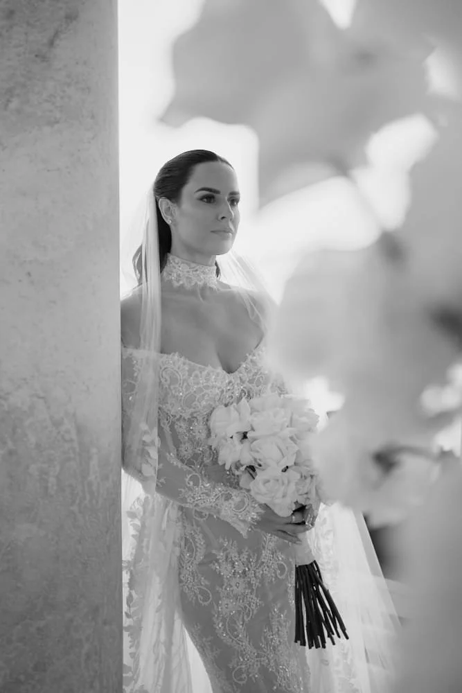A bride in a lace wedding dress holding a bouquet of flowers, standing by a wall with a blurred background of leaves.