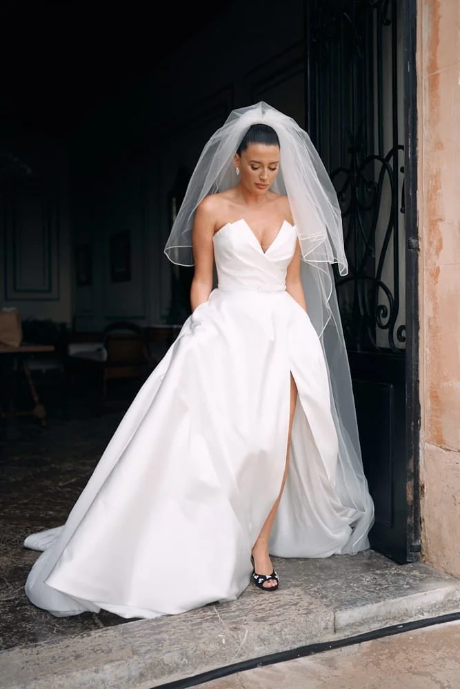 Bride in a white wedding gown with a thigh-high slit, standing on a stone floor by a doorway, wearing a veil and black open-toe heels.