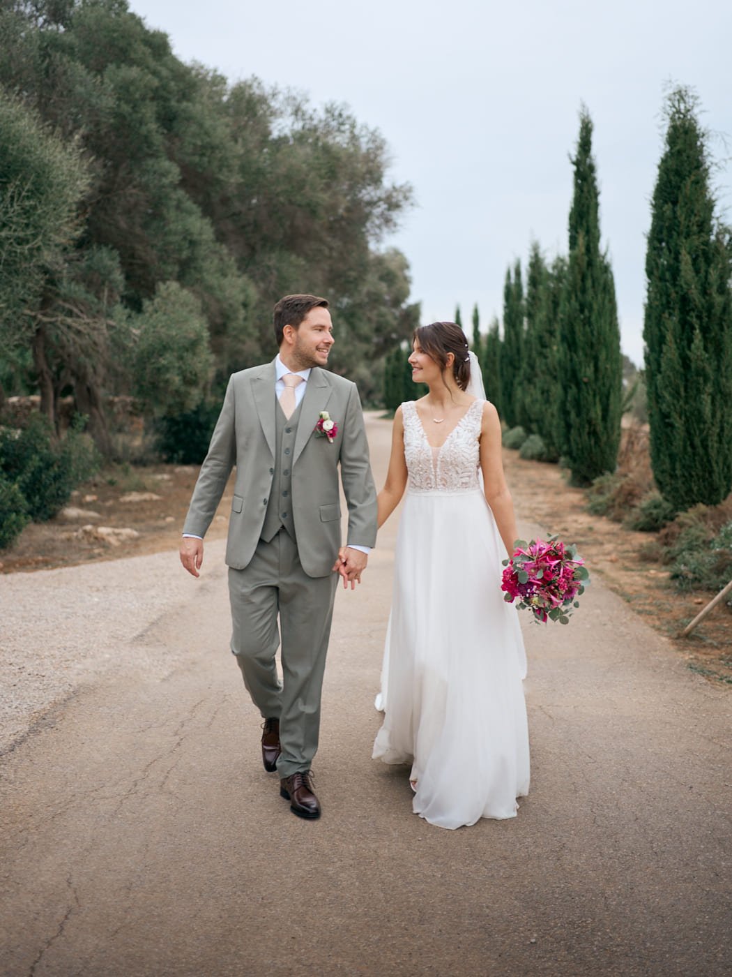 A bride and groom walking hand in hand outdoors, smiling at each other, with trees and a cloudy sky in the background. Wedding in Mallorca. The Lumery.