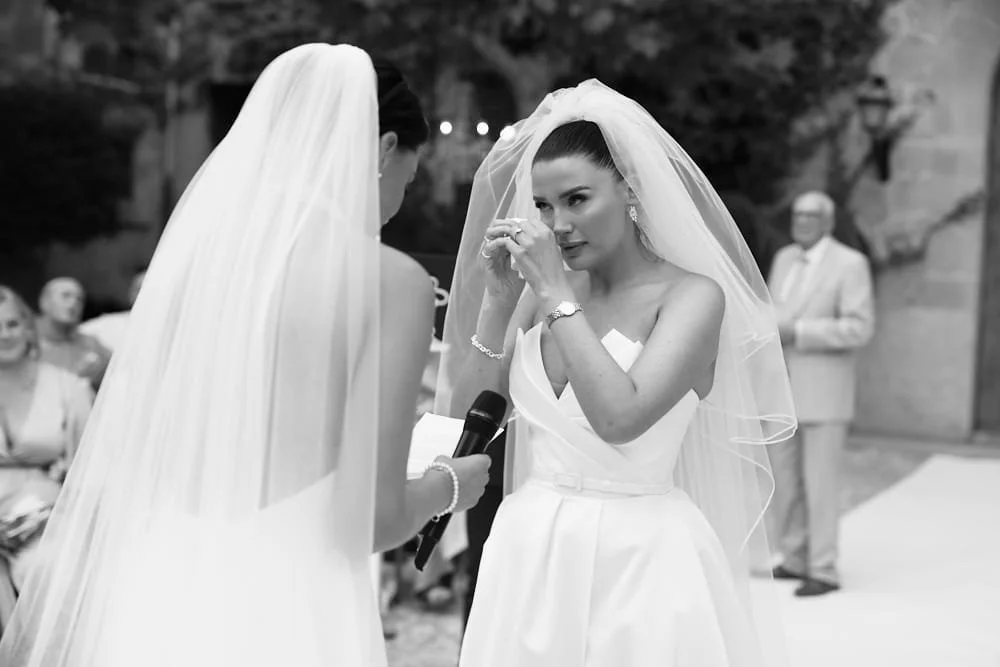 A bride in a wedding dress and veil tears up during her wedding ceremony as the officiant reads vows, while guests watch in the background.