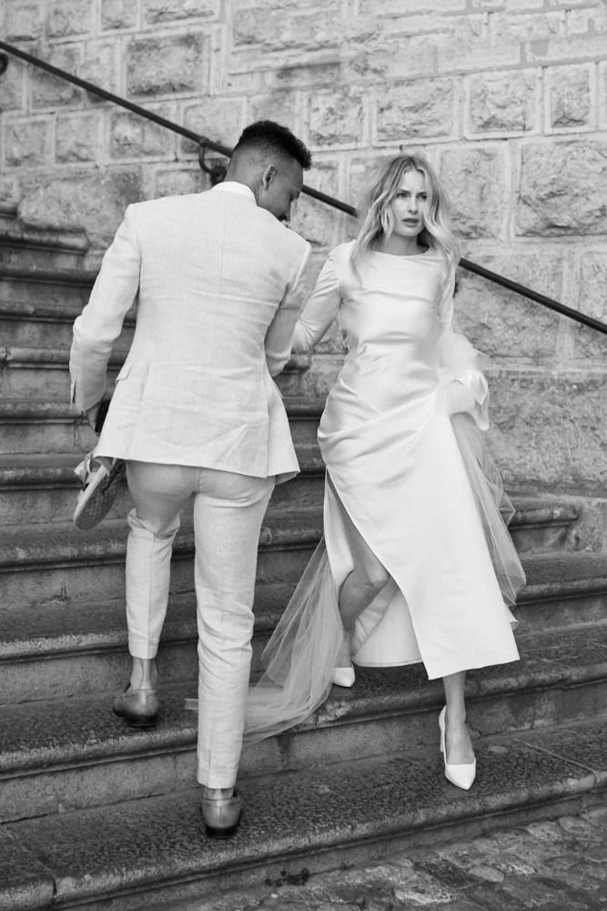 A woman in a wedding dress holding her dress while walking down stairs, assisted by a man in a light-colored suit, with an urban stone building background.
