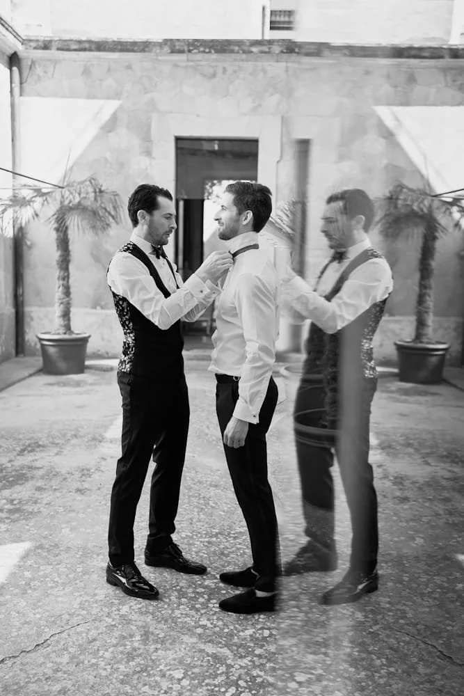 Two men dressed in formal attire in a courtyard, with one adjusting the other's bow tie while a ghostly mirror image of the scene appears to the right.