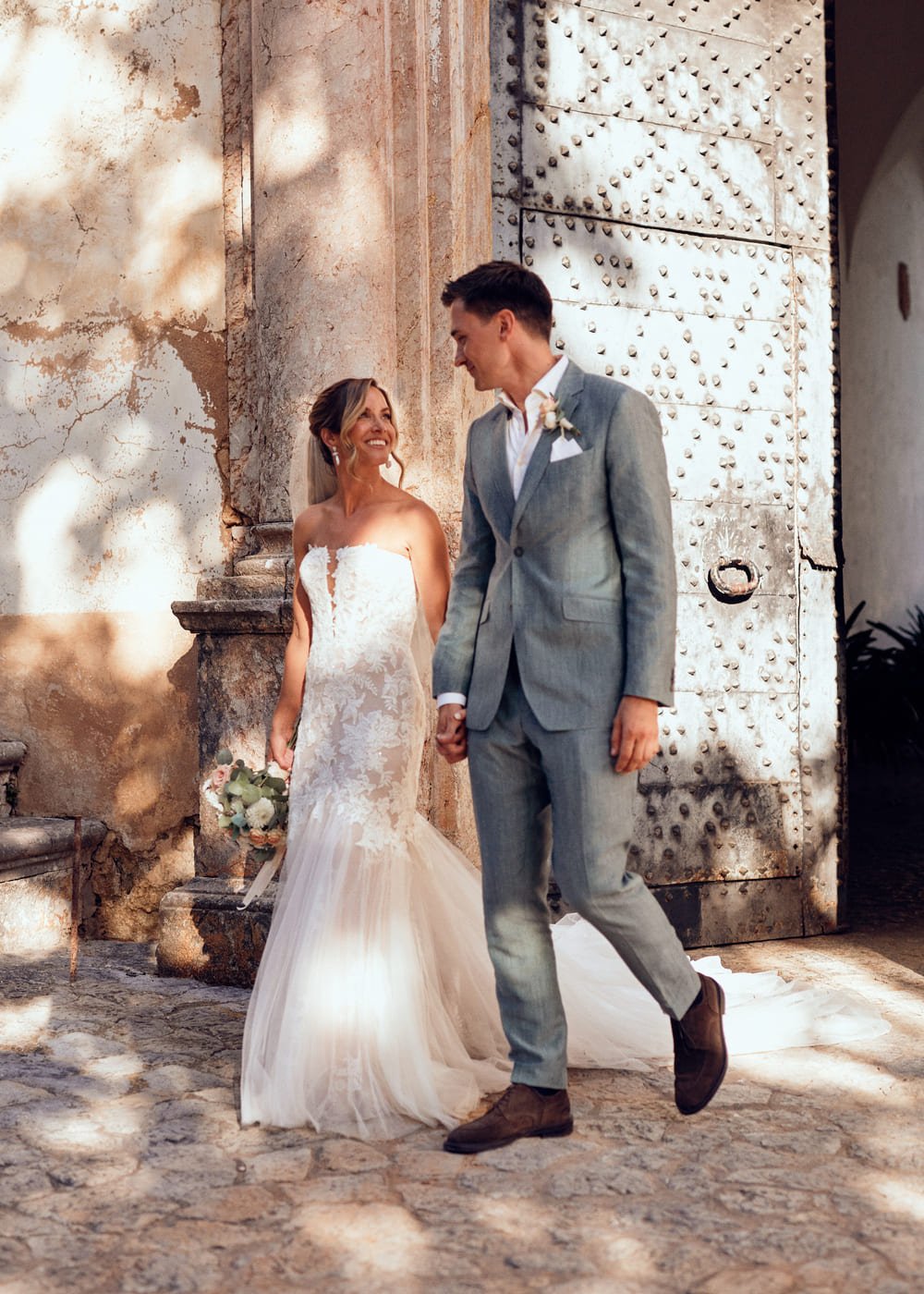 Bride and groom smiling at each other in front of an old stone building, bride holding a bouquet.