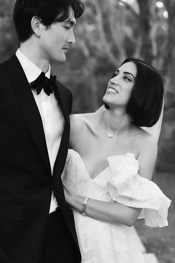 A black-and-white photo of a man and woman dressed in formal wedding attire, looking at each other outdoors with trees in the background.