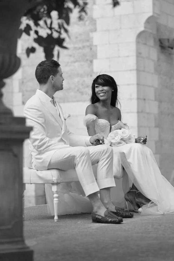 A bride and groom sitting on a couch during their wedding ceremony, holding hands and smiling at each other.