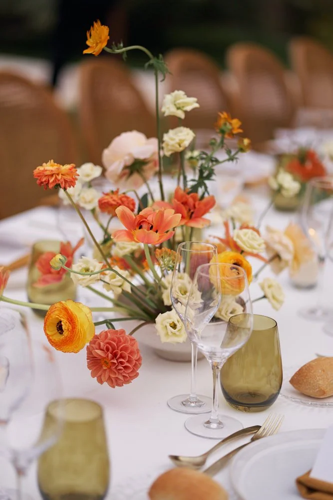 A table decorated with colorful flowers in a vase, with wine glasses, plates, and gold cutlery set for a meal, and bread rolls on the table.