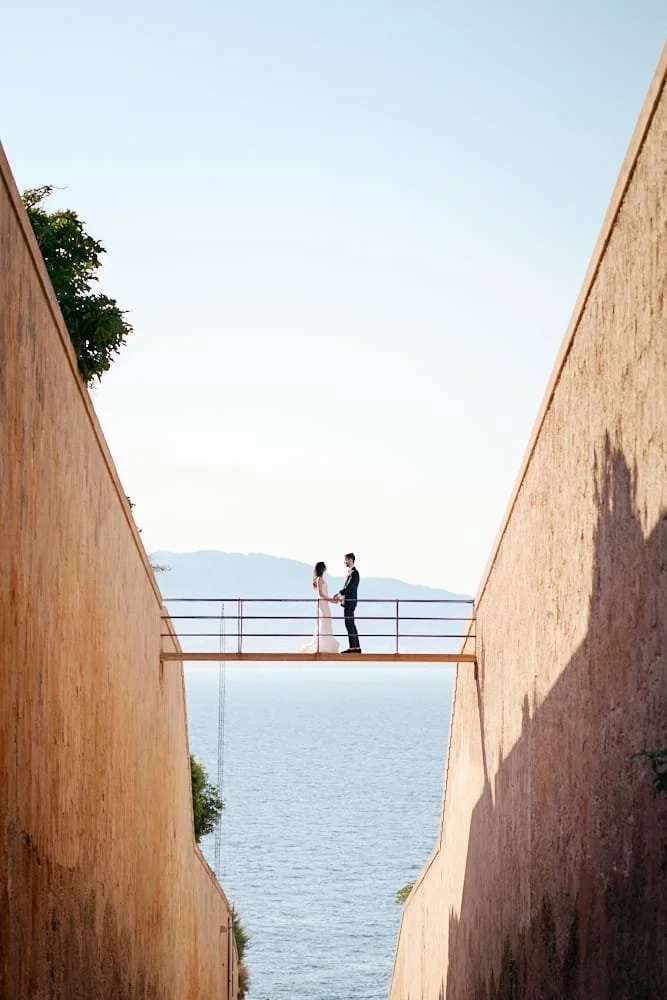 A couple standing on a narrow bridge between two tall brick walls above a body of water, holding hands and facing each other.