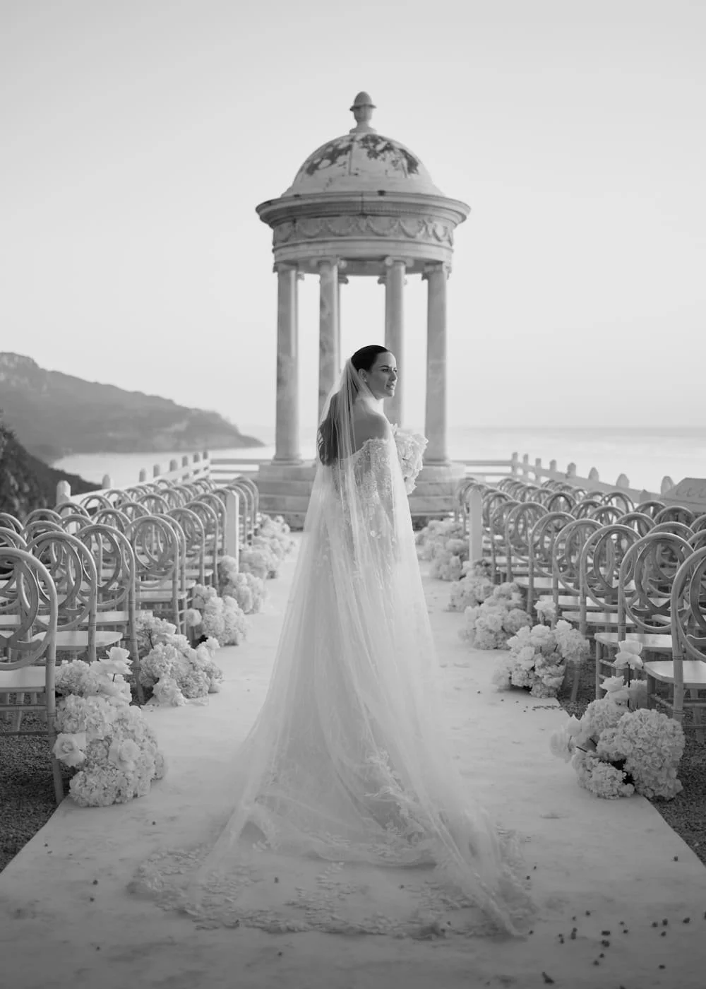 A bride in a wedding dress with a long train stands on an outdoor wedding aisle decorated with flowers, facing a decorative pavilion, with a scenic coastal view in the background. Son Marroig.