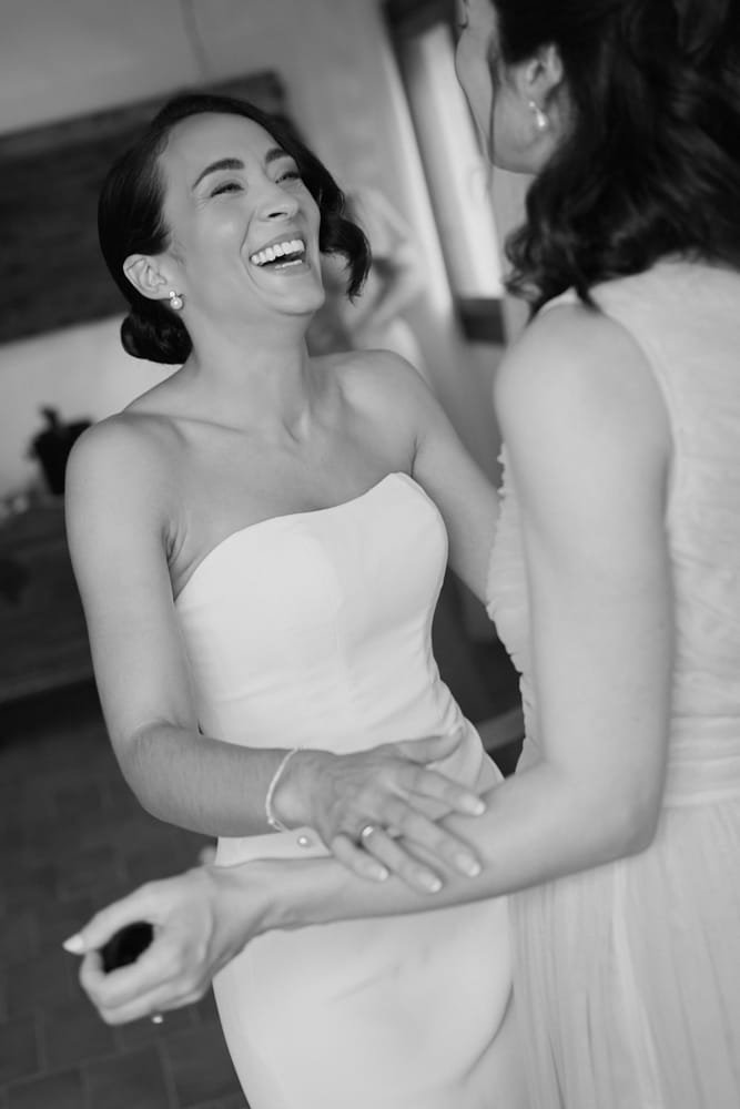 Two women, one in a white strapless dress, are holding hands and smiling joyfully, likely at a wedding or celebration.