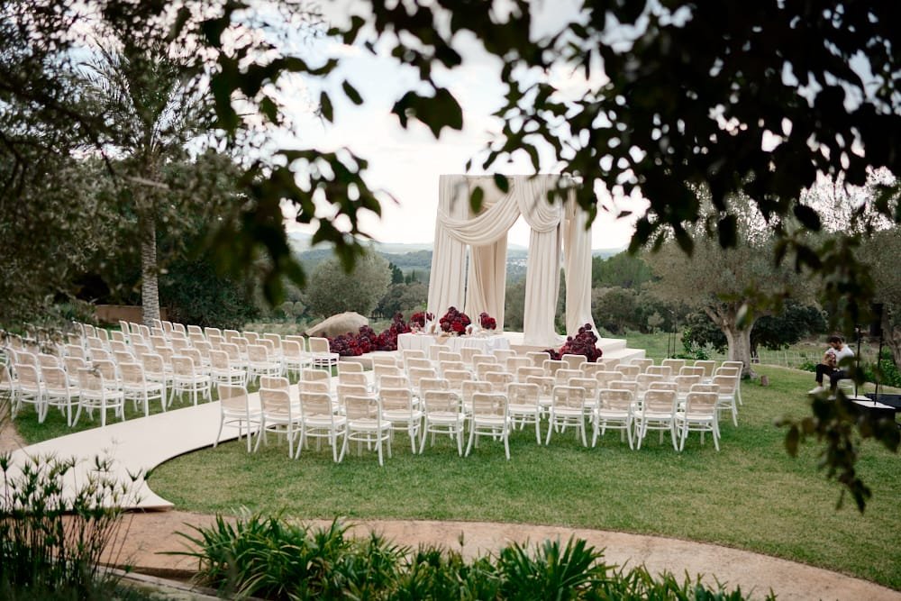Outdoor wedding ceremony setup with white chairs, draped fabric backdrop, and rose centerpieces, surrounded by trees and greenery.