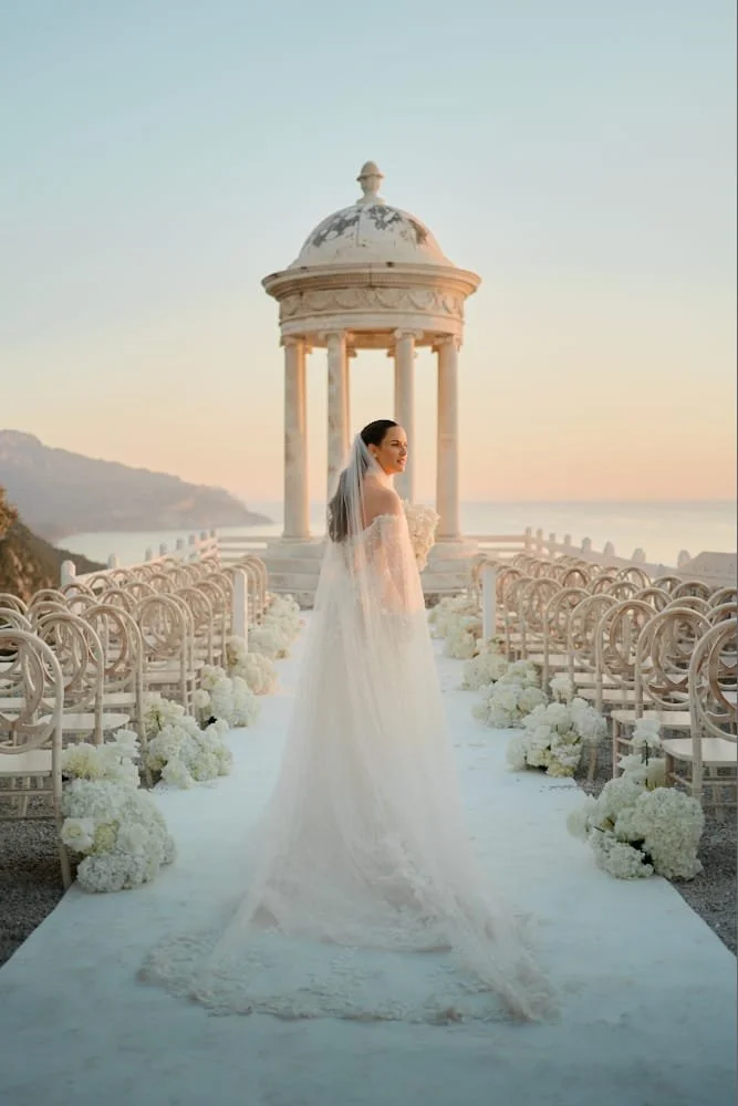 Bride in a wedding gown with a long train and veil standing on an outdoor wedding aisle decorated with white flowers, with a classical pavilion and scenic ocean view in the background during sunset.