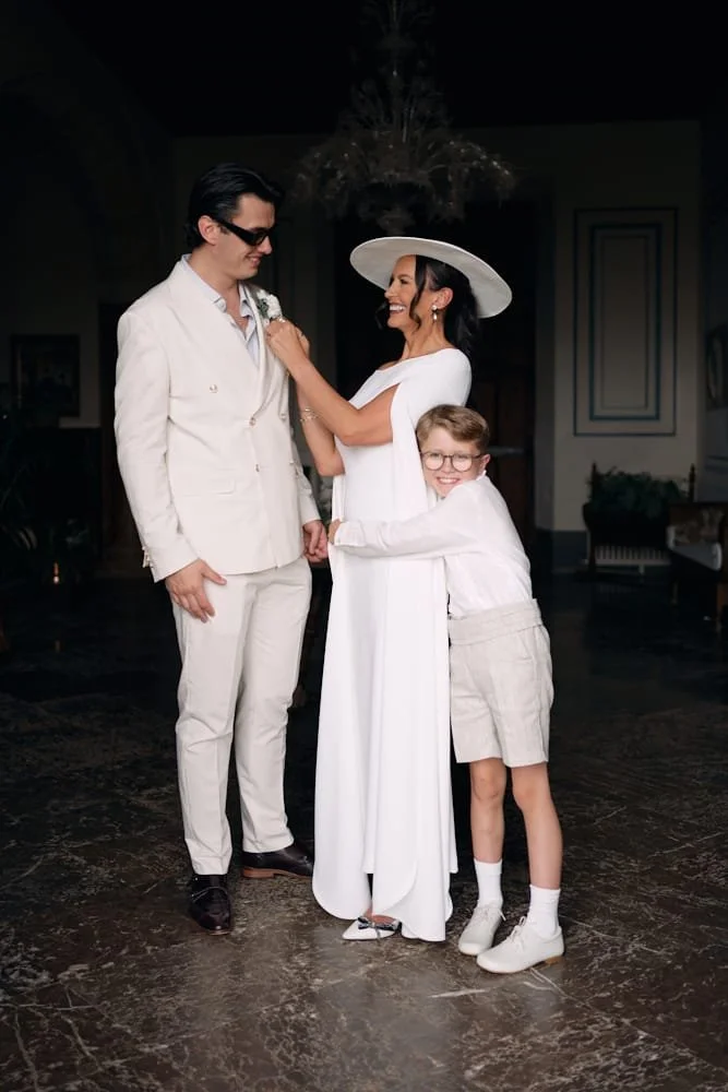 A woman in a long white dress and wide-brimmed hat, smiling with a young boy in glasses and shorts hugging her, standing with a man in a white suit in an elegant indoor setting with a chandelier.