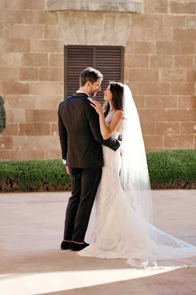 A bride and groom stand close together outdoors, gazing into each other's eyes during their wedding. The bride wears a white wedding dress with a long train and veil. The groom is dressed in a black tuxedo. Behind them is a brick building with a wind