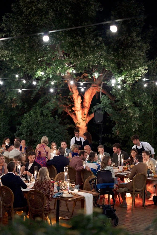 A nighttime outdoor dining event with guests seated at tables under string lights and a large tree, with waitstaff serving food.