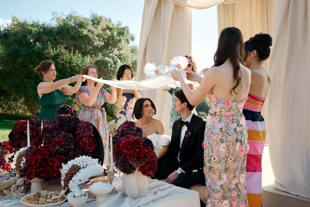 A wedding ceremony in a garden with brides and grooms seated, surrounded by women holding a cloth or veil over them, with flowers and candles on a table in front.