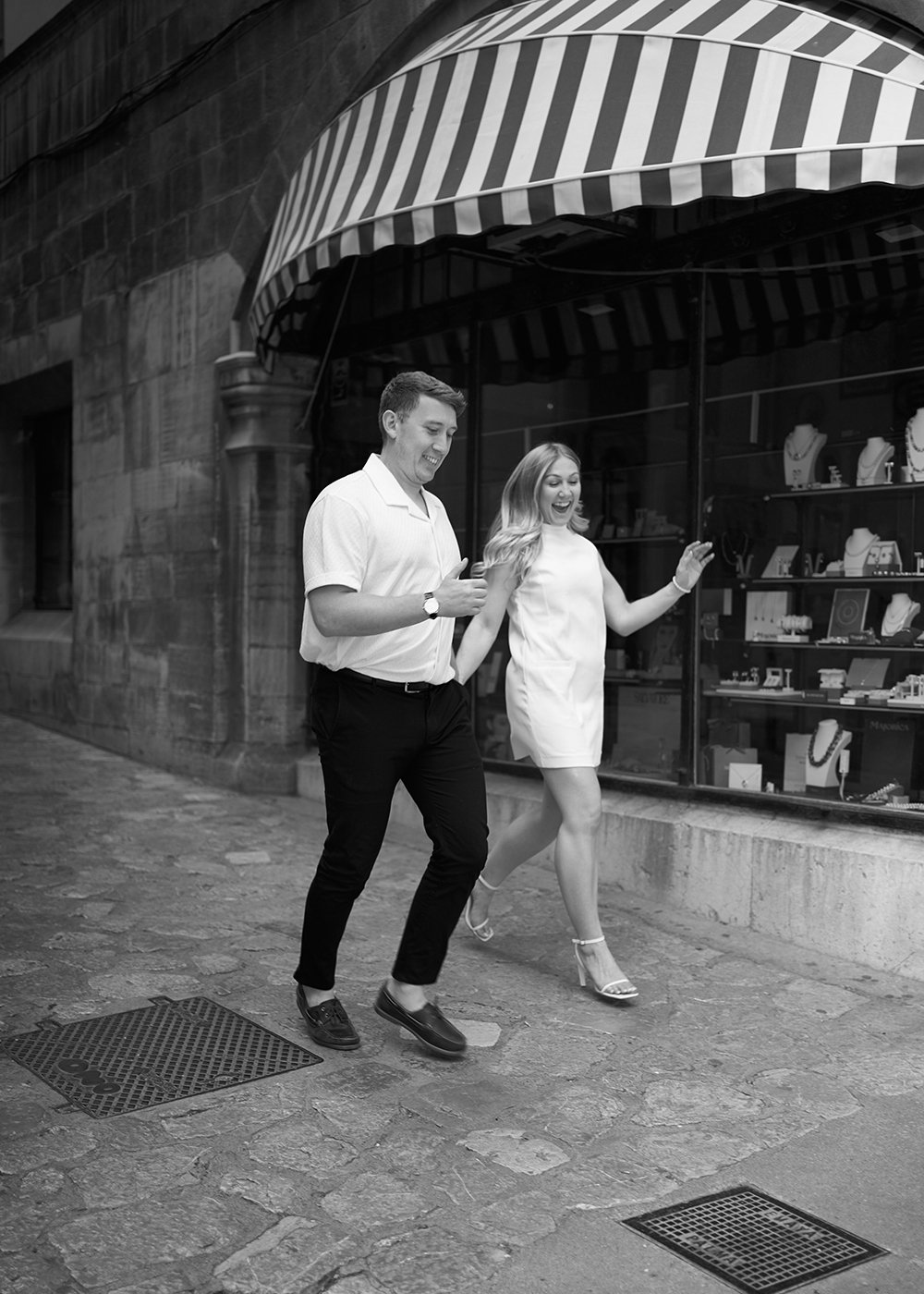 Black and white photo of a man and woman walking joyfully on a stone-paved street, in front of a shop with striped awning and window display of jewelry.